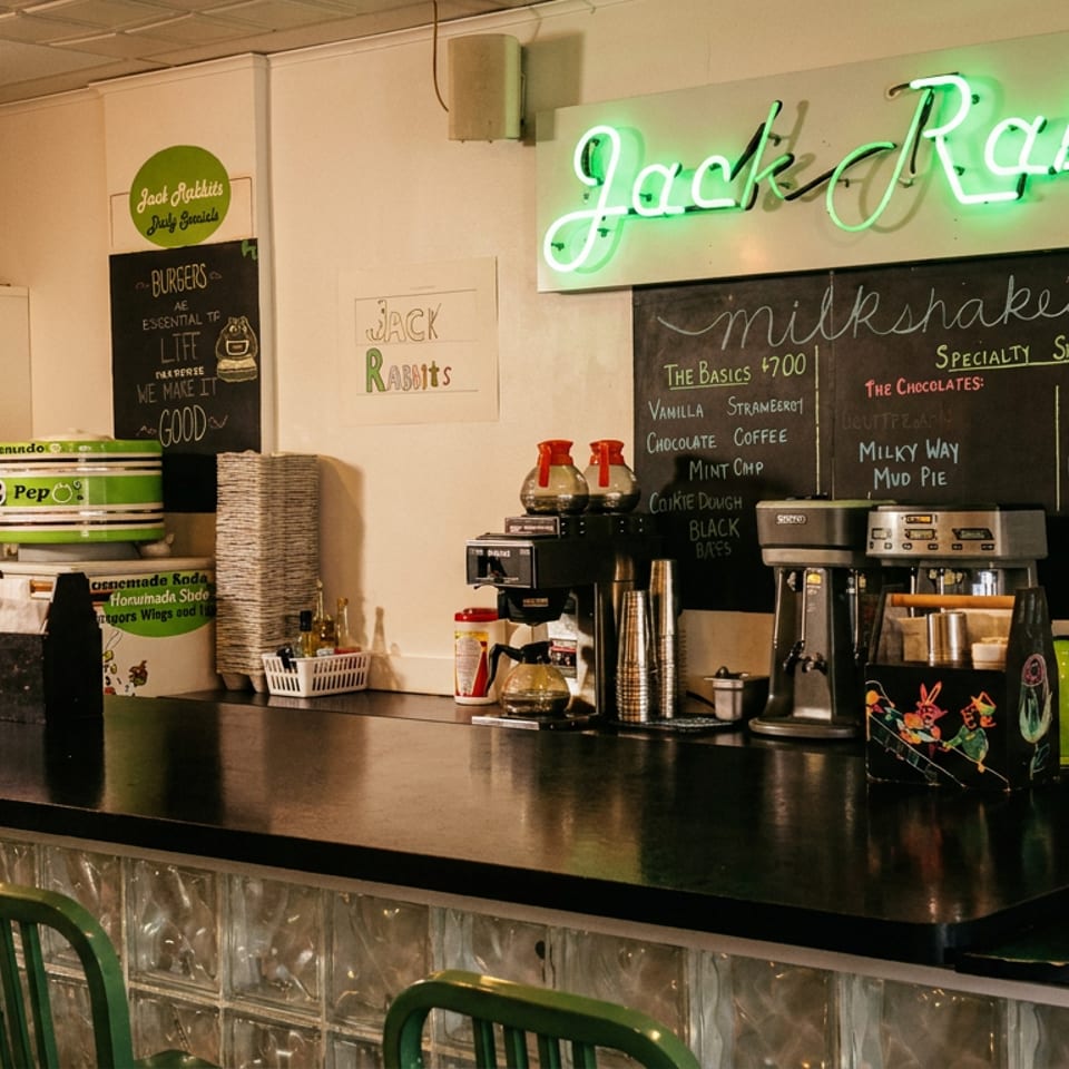 A diner counter with green bar stools, coffee machines, and a neon "Jack Rabbit's" sign.