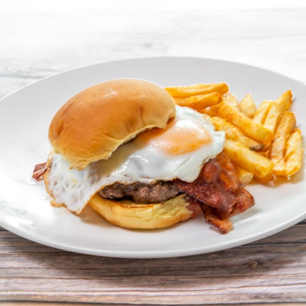 Bacon cheeseburger topped with a fried egg, served with golden fries on a white plate.