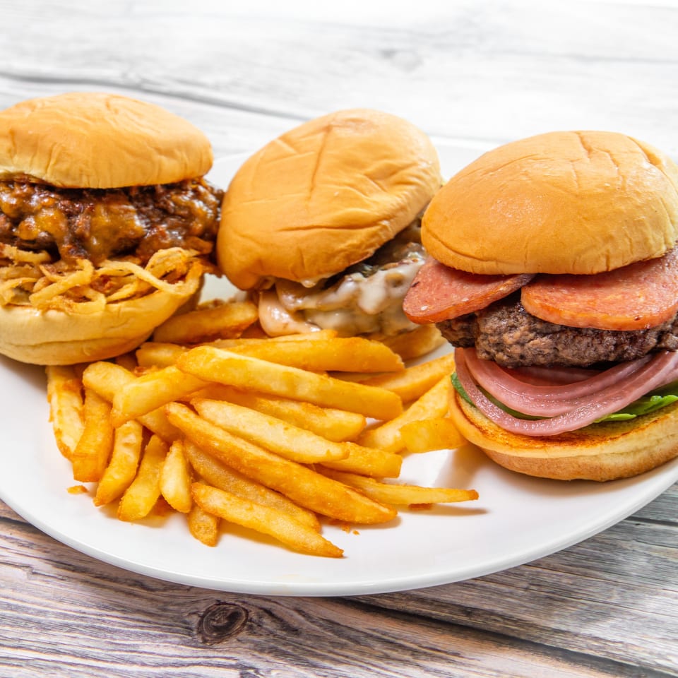 Three loaded burgers, including bacon and cheeseburgers, served with crispy French fries on a white plate.
