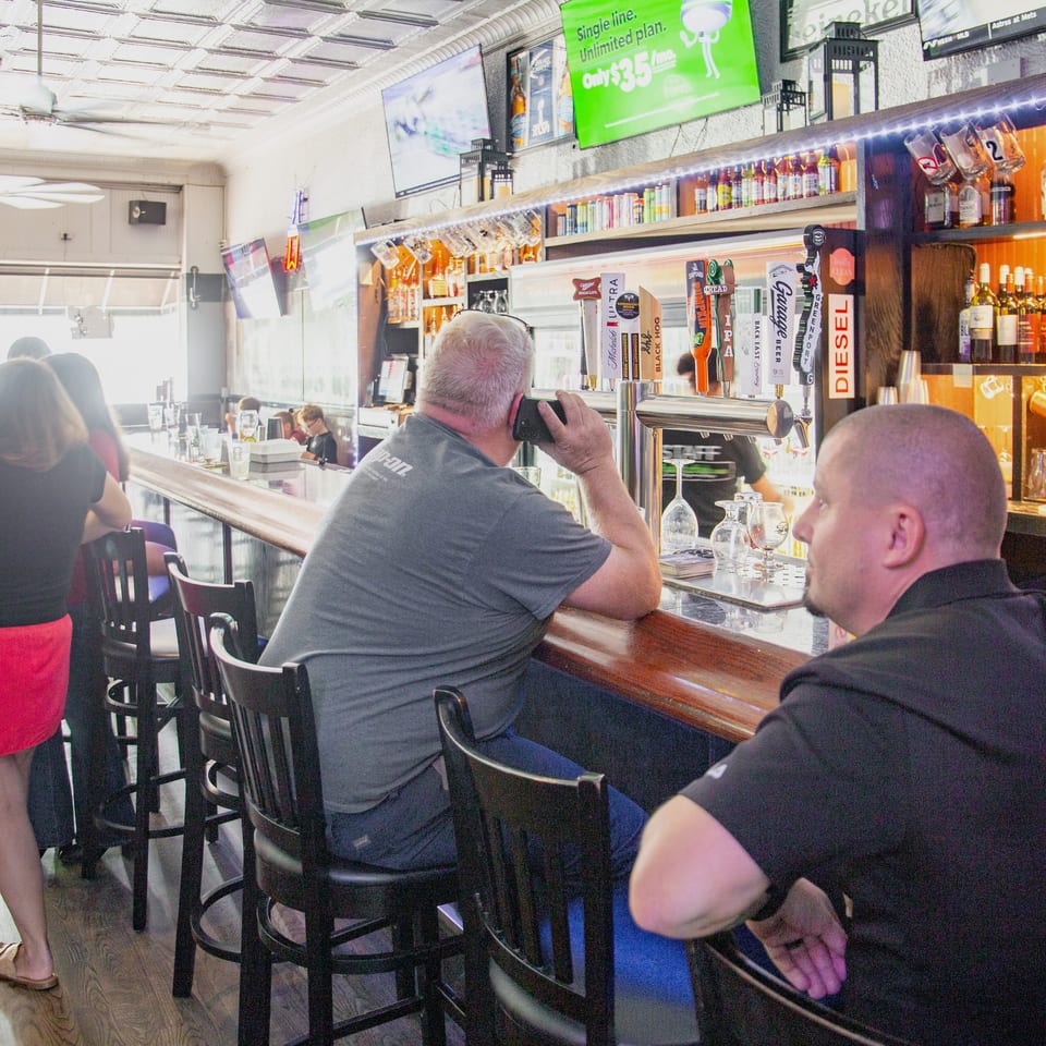 Busy sports bar with patrons seated at the counter, illuminated liquor shelves, beer taps, televisions, and baseball-themed décor.