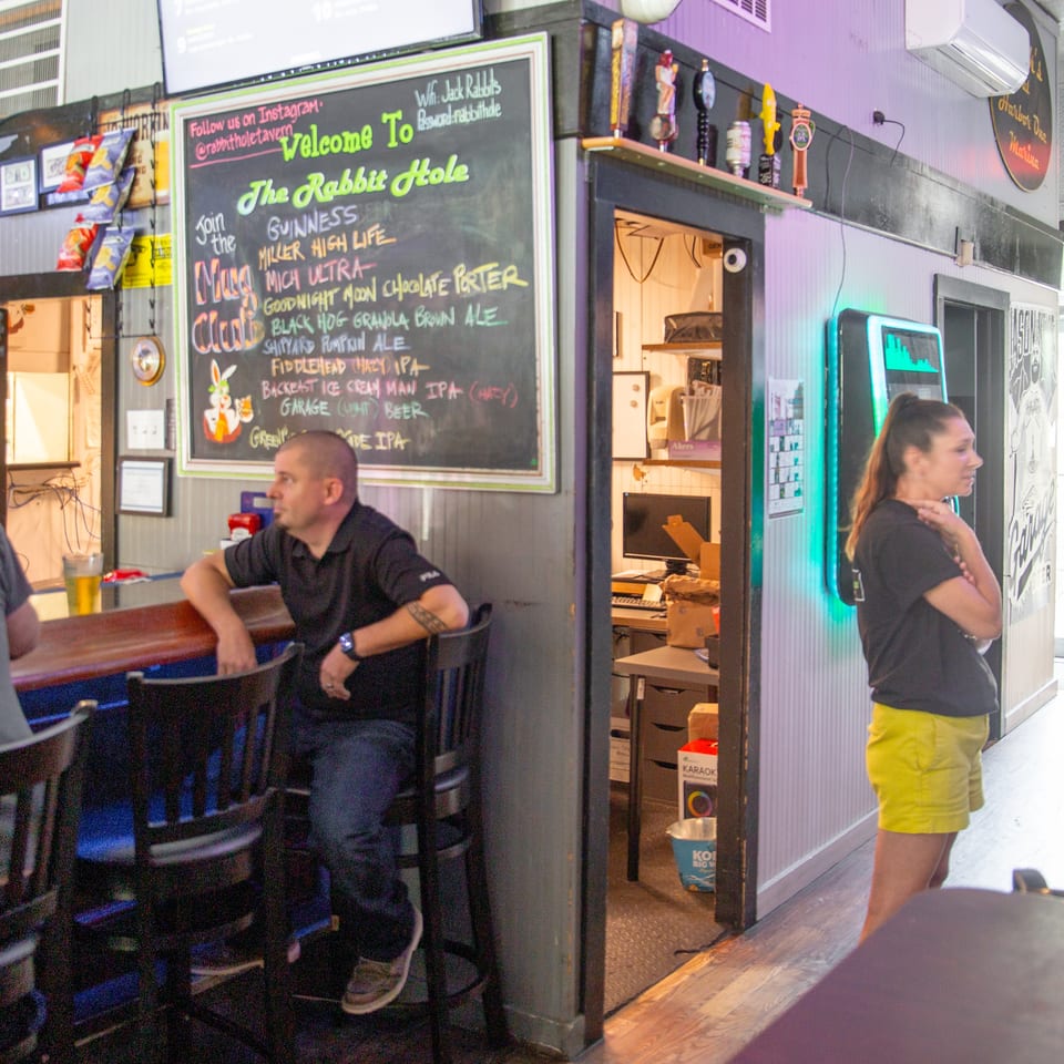 Inside The Rabbit Hole bar, patrons sit at the counter beside a chalkboard beer list, beer taps, and colorful wall décor.
