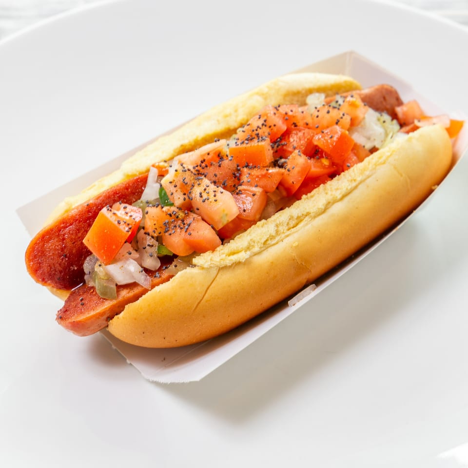 Loaded hot dog in a toasted bun topped with diced tomatoes, onions, peppers, and black poppy seeds.