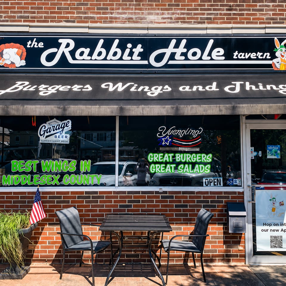 The Rabbit Hole Tavern storefront with an awning, outdoor seating, and window signs for burgers and wings.