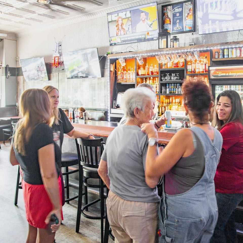 Patrons gather at a lively restaurant bar with liquor shelves, beer refrigerators, televisions, and high-top tables.
