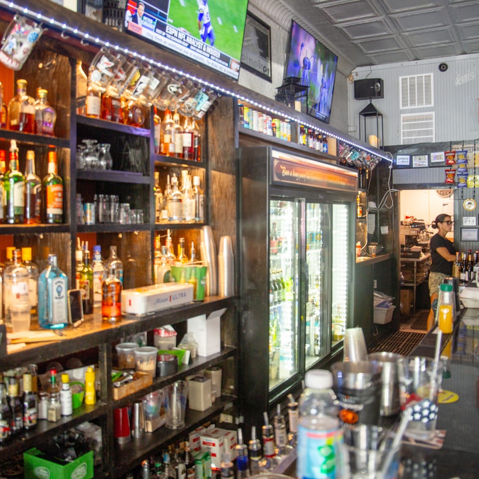Busy restaurant bar lined with liquor bottles, a beverage cooler, sports televisions, and a bartender working behind the counter.
