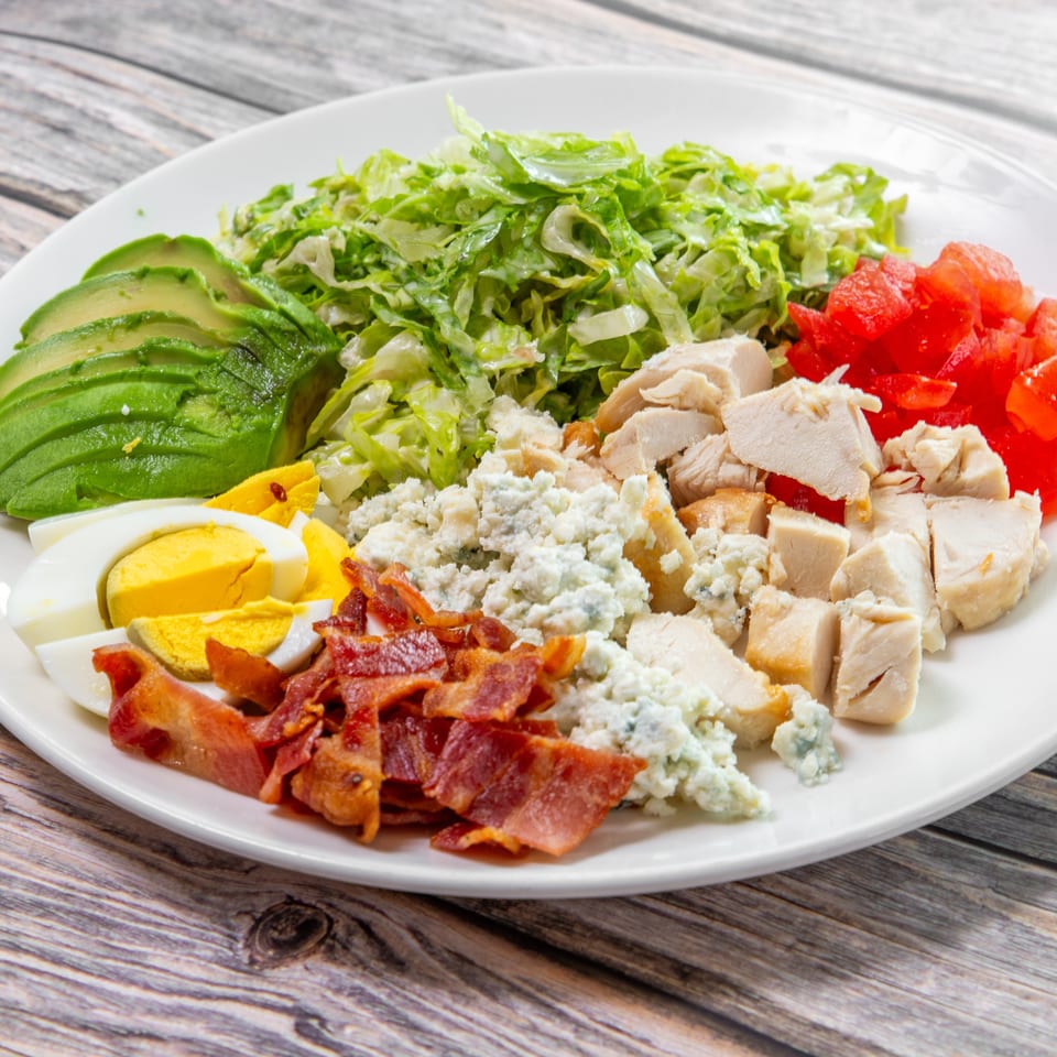 Cobb salad with lettuce, grilled chicken, avocado, tomatoes, boiled eggs, bacon, and blue cheese on a white plate.