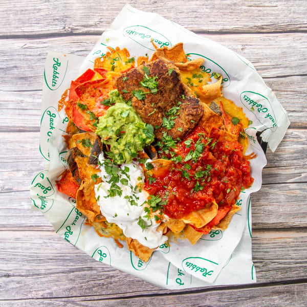 Loaded nachos topped with grilled meat, melted cheese, guacamole, salsa, sour cream, and cilantro on branded paper.
