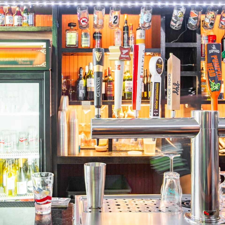 Brightly lit restaurant bar with numerous beer taps, liquor bottles, glassware, and a refrigerated drink display.