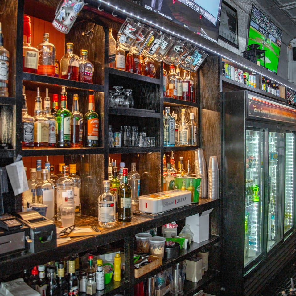 Well-stocked bar displays liquor bottles, mixers, glassware, a refrigerated drink case, and sports playing on overhead televisions.