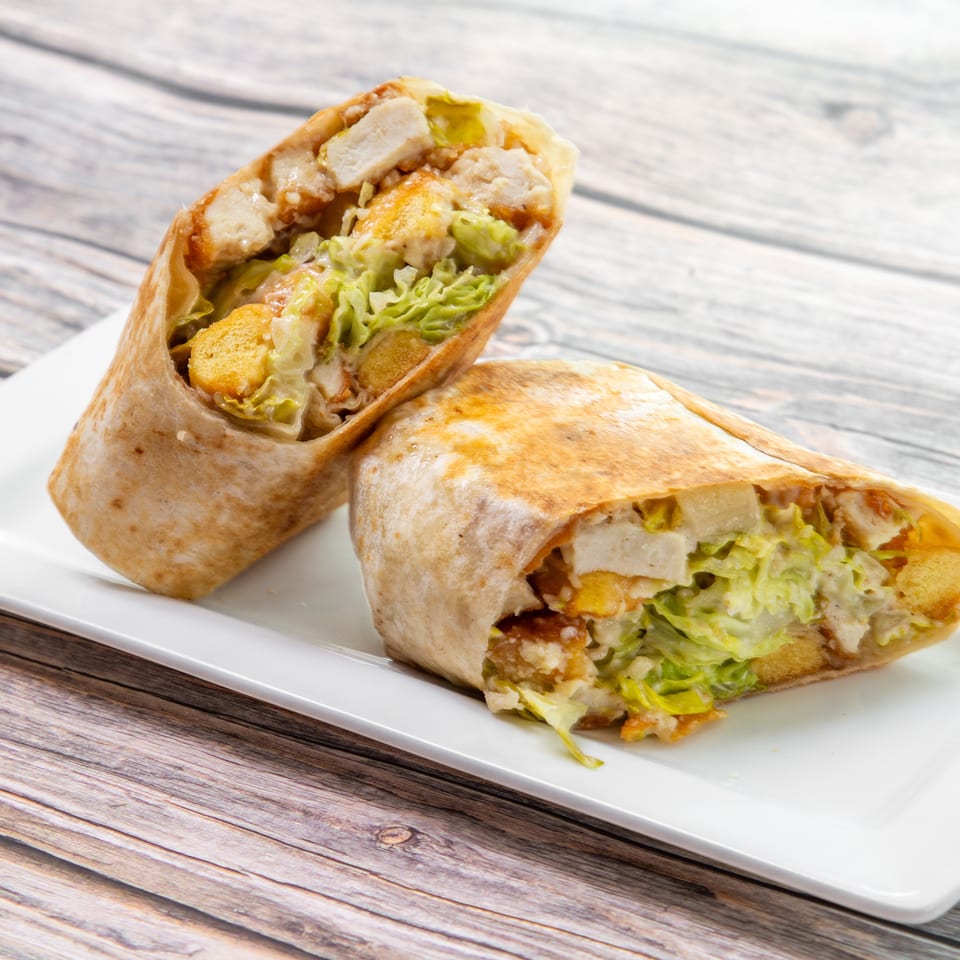 Two toasted chicken wraps filled with lettuce and crispy pieces, sliced open on a white plate over a wooden table.