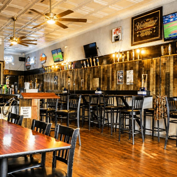 A rustic bar interior with wooden walls, high-top tables, bar stools, and TVs showing sports.