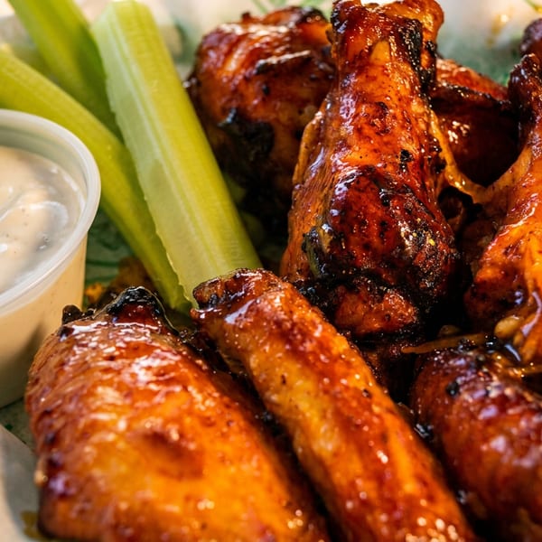 Close-up of a basket of glossy, barbecued chicken wings with celery sticks and a side of ranch.