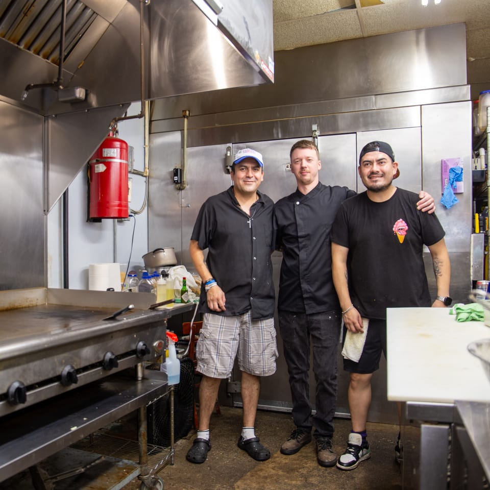 Three restaurant staff members pose together in a commercial kitchen beside a large grill and stainless-steel equipment.