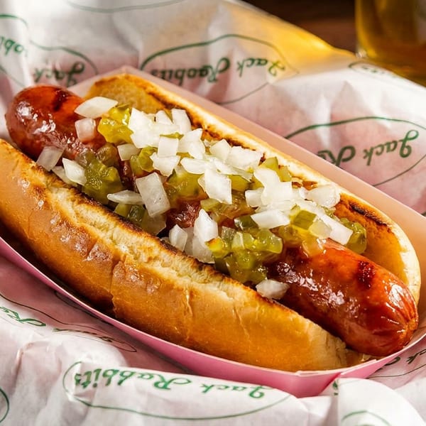 A Jack Rabbits hot dog with relish and diced onions in a paper tray, with a drink in the background.