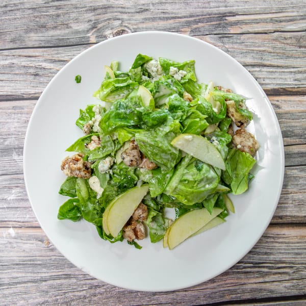 Green lettuce salad with sliced apples, blue cheese, crispy toppings, and creamy dressing on a white plate.
