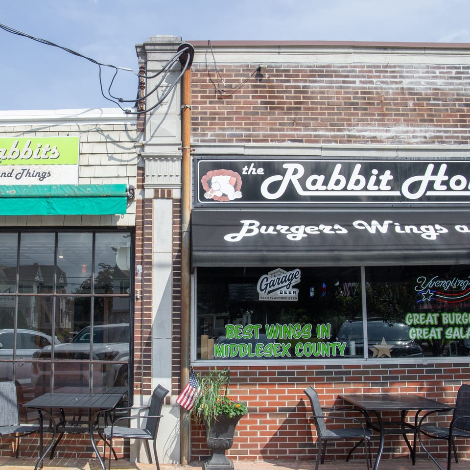 The Rabbit Hole restaurant exterior features brick walls, bold signage, outdoor seating, and windows advertising burgers, wings, and salads.