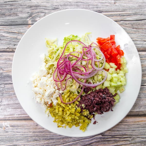 Overhead view of a colorful chopped salad with lettuce, tomatoes, cucumber, red onion, feta, olives, and peppers on a white plate.