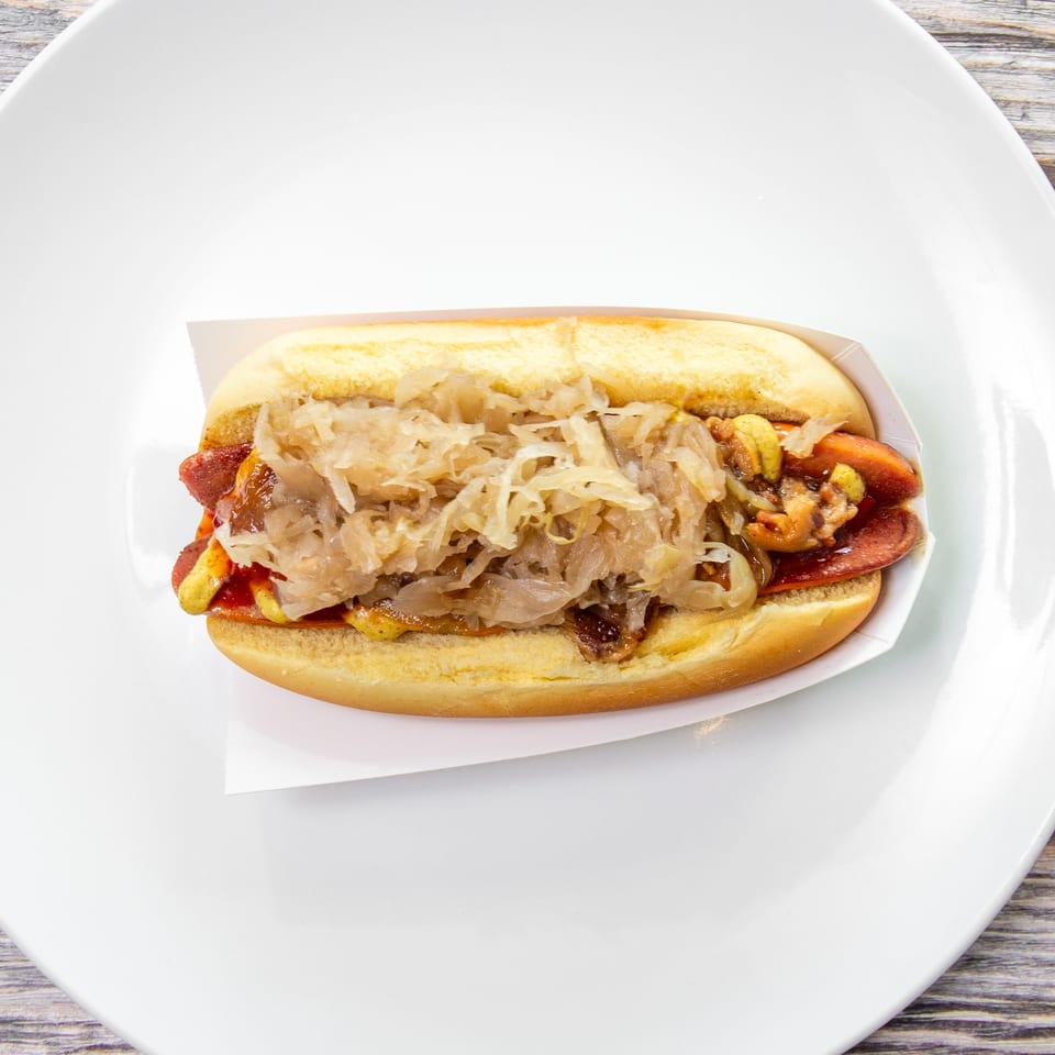 Loaded hot dog topped with sauerkraut, mustard, relish, and onions in a bun on a white plate.